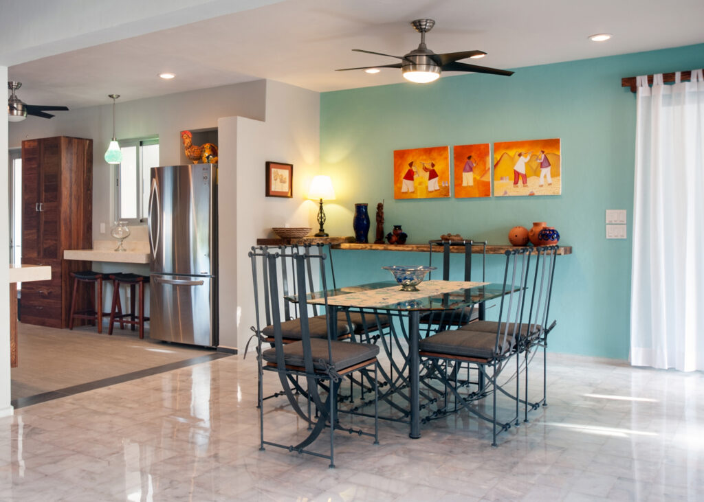 Dining table with black chairs beside a turquoise accent wall, with the kitchen behind.