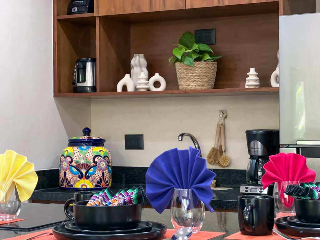 Kitchen sink and coffee maker beneath wooden shelves holding ceramics and a potted plant.