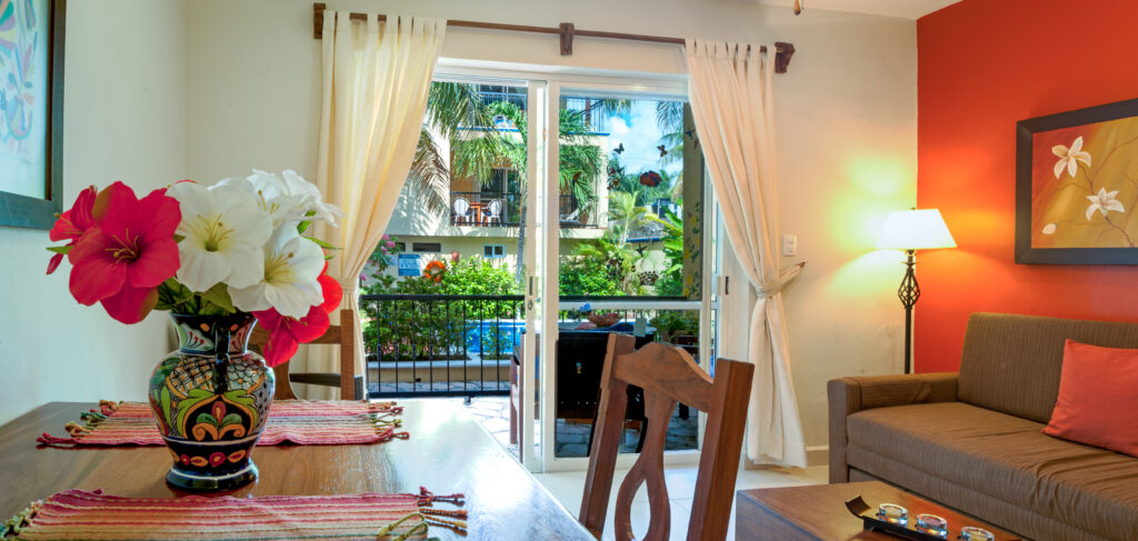 Flowers on a wooden dining table beside a sofa and glass doors overlooking palms.