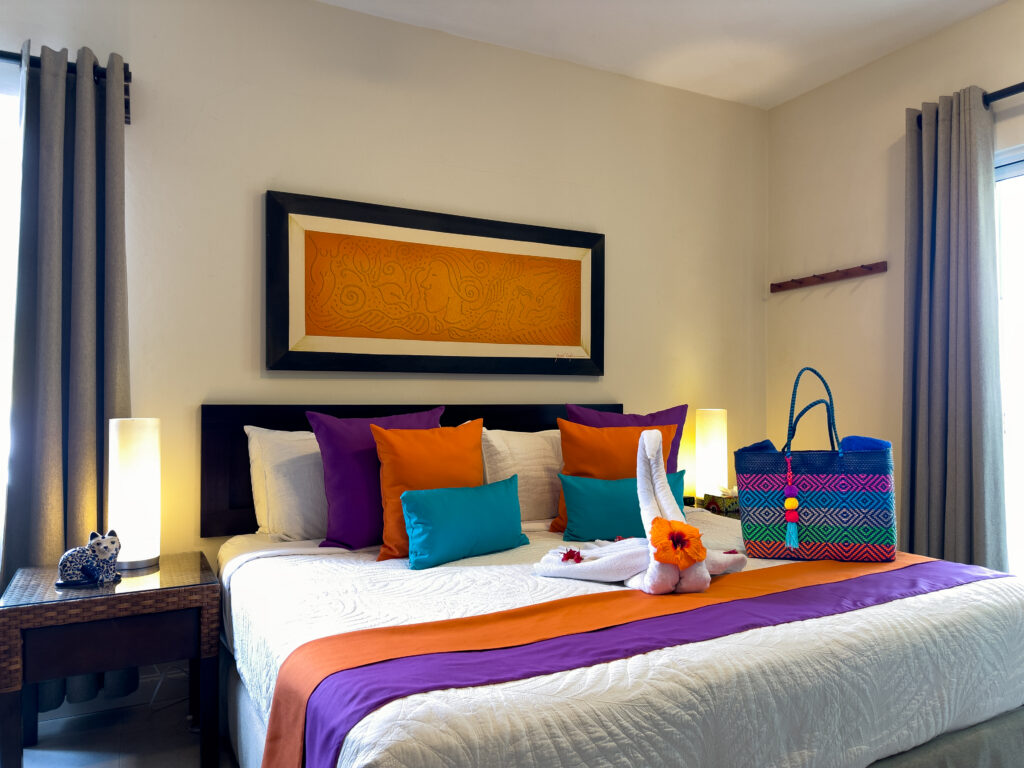 Bed with orange and purple runners, colorful pillows, and a towel swan beneath framed artwork.