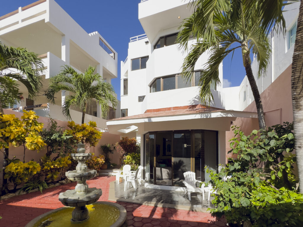 Tiered fountain in a planted courtyard in front of a white multistory house with balconies.