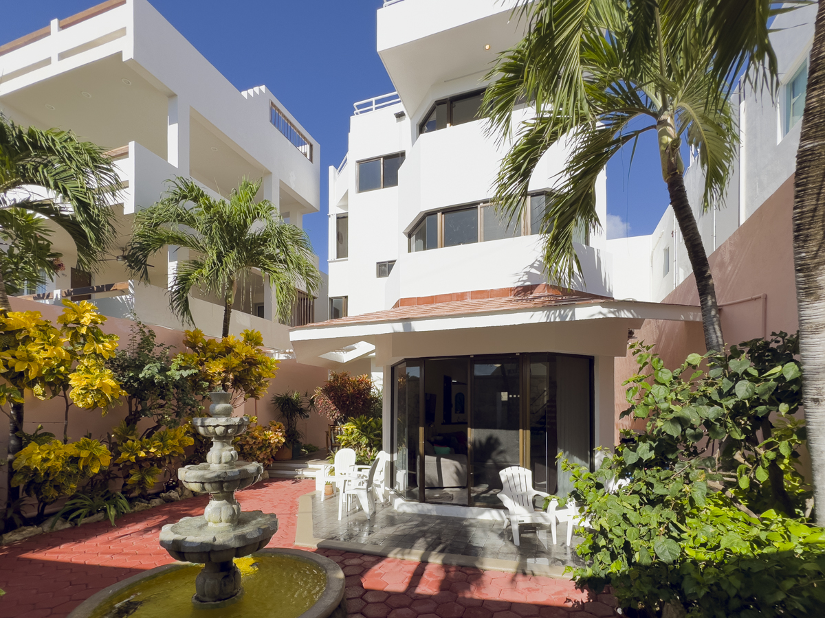Tiered fountain in a planted courtyard in front of a white multistory house with balconies.