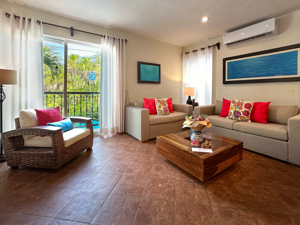 Sofas and a wicker chair around a wooden coffee table beside glass doors overlooking palms.