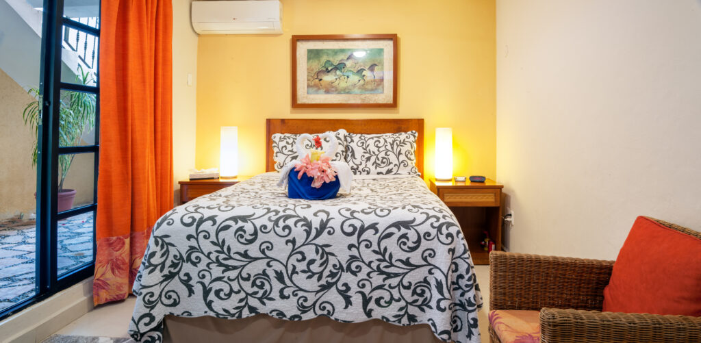 Bed against a yellow accent wall with matching bedside lamps and an orange-cushioned wicker chair.