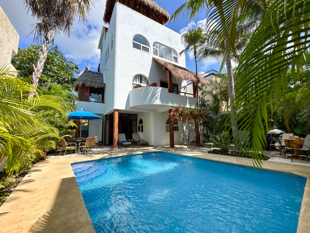 Blue pool surrounded by palms beneath a white multistory building with thatched awnings.