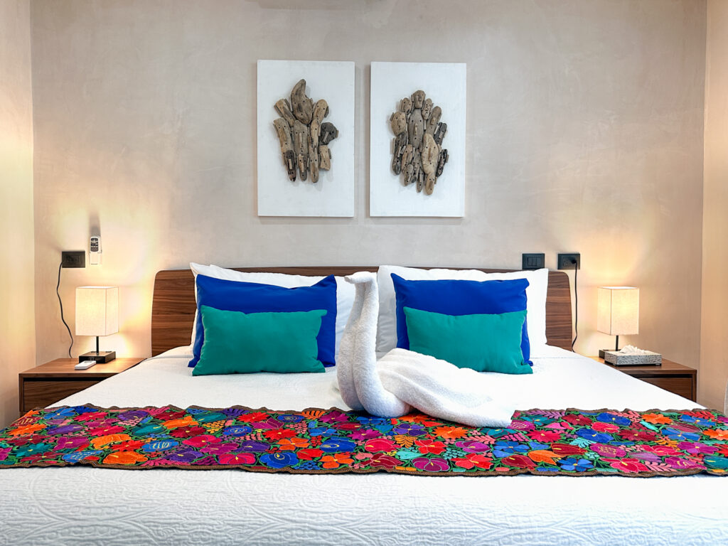 White bed with blue and turquoise pillows and a towel swan on a floral runner.