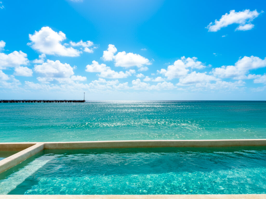 Pool water in the foreground with turquoise sea and a distant pier under scattered clouds.