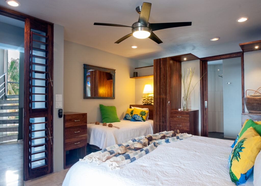 Larger bed and a single bed with matching tropical cushions beneath a ceiling fan.