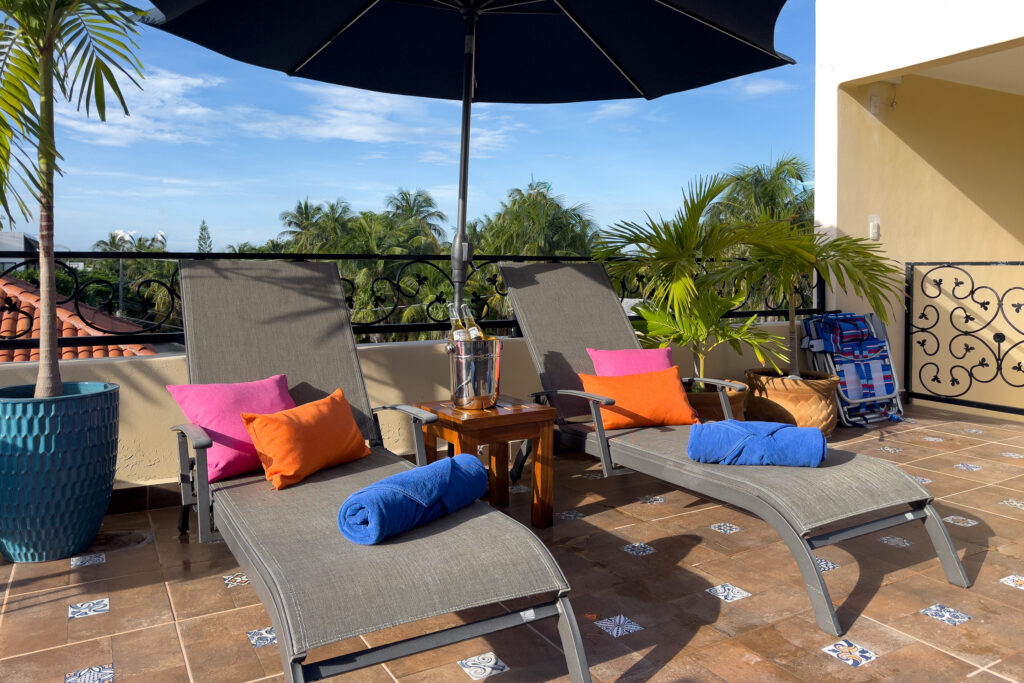 Two terrace lounge chairs with bright cushions and rolled blue towels beneath a dark umbrella.
