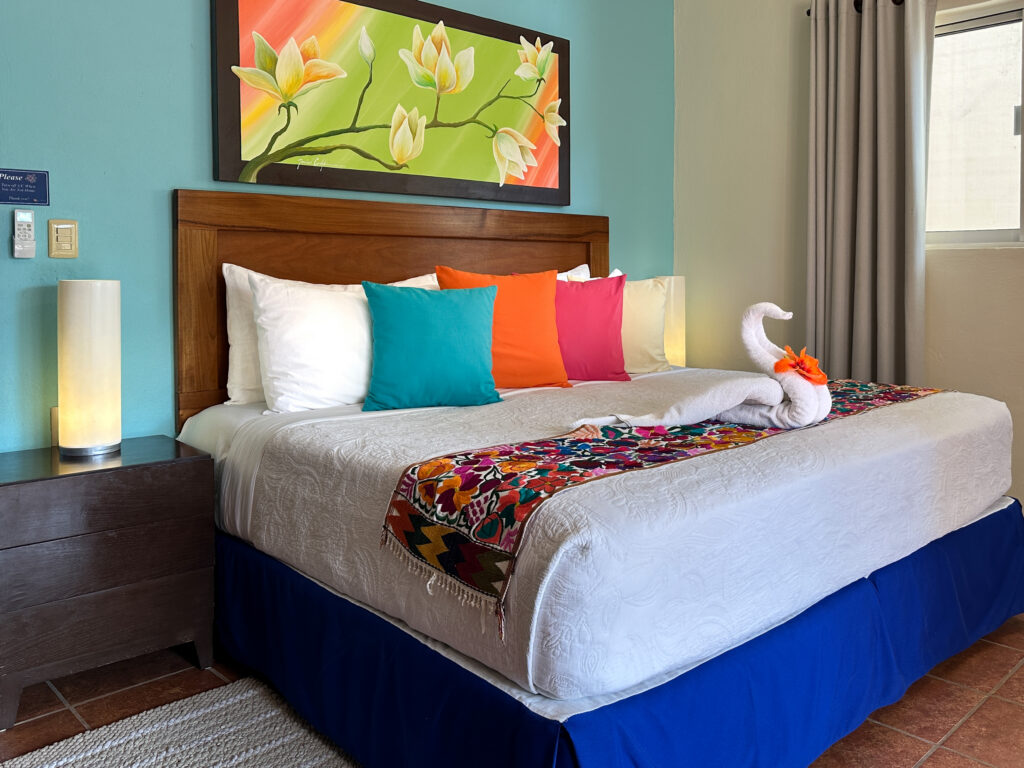 White bed with bright pillows, a floral runner, and towel swan beneath green floral artwork.
