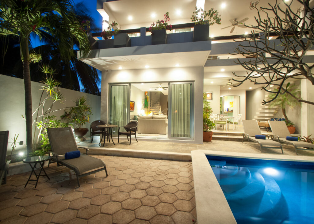 Illuminated pool and patio seating outside open glass doors at night.