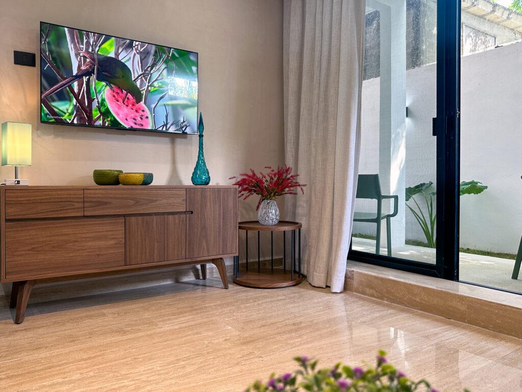 Wall-mounted television above a wooden console beside a floor vase of flowers and glass patio doors.