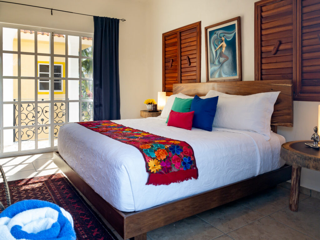 White bed with bright pillows and a floral runner beside French doors and wooden shutters.