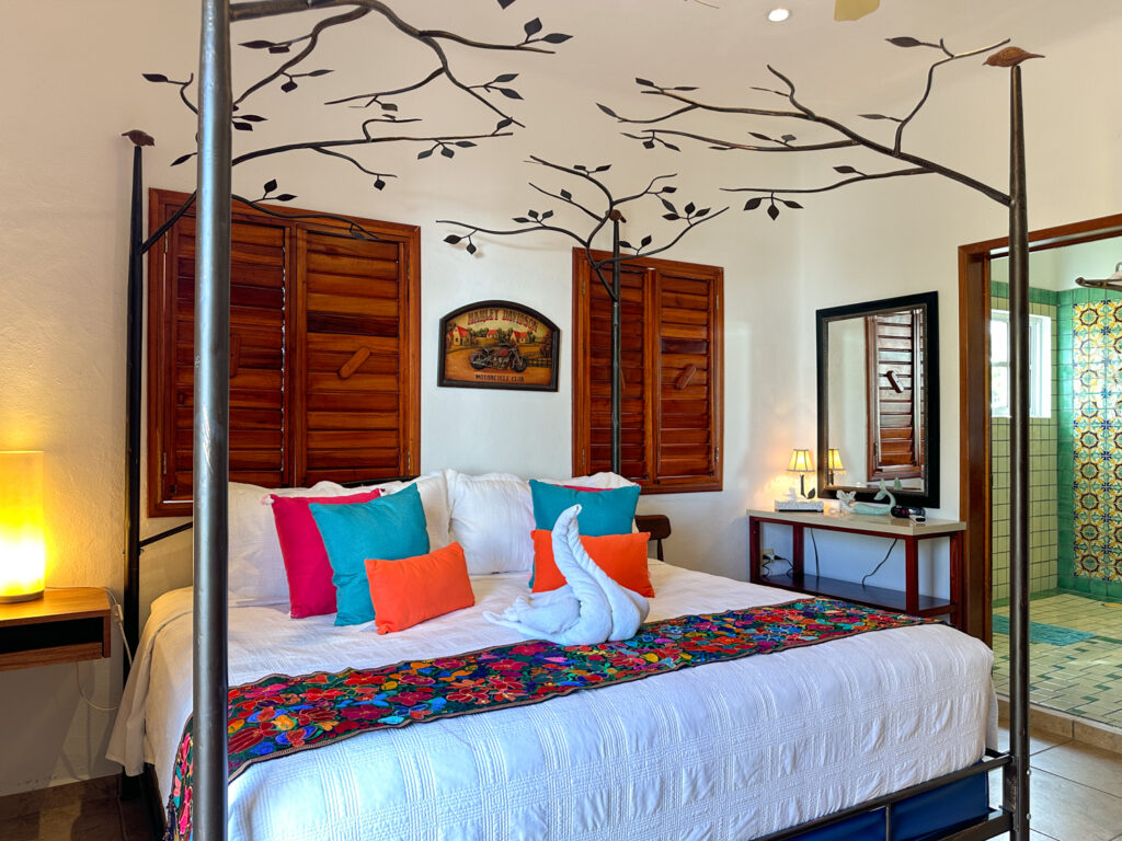 Four-poster bed with bright cushions and towel swan beneath a painted branch mural and wooden shutters.