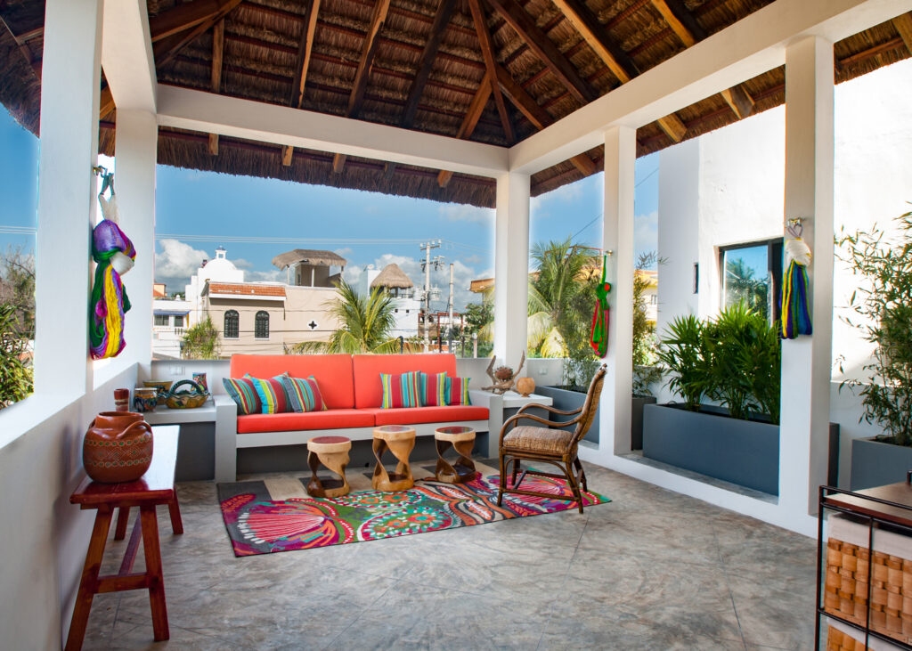 Orange-cushioned bench, wicker chairs, and colorful rug beneath a high thatched terrace roof.