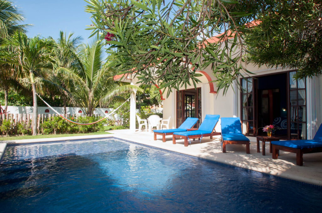 Swimming pool shaded by leafy branches beside lounge chairs and open house doors.