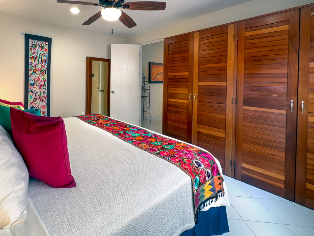 Wooden closet doors beside a bed with colorful bedding and an open bathroom doorway.