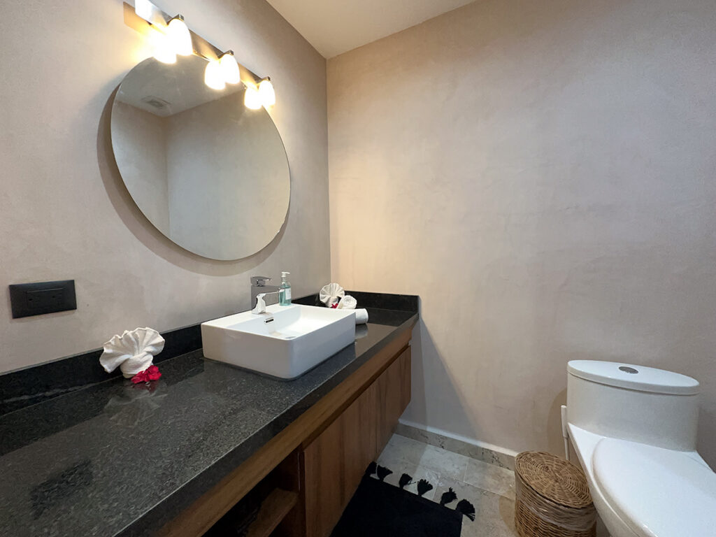 Rectangular white basin on a dark counter beneath a round mirror beside a toilet.