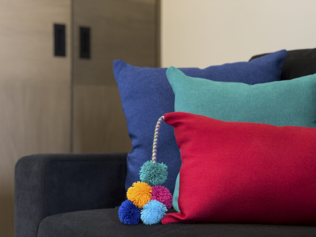 Red, turquoise, and blue cushions with pom-pom trim on a dark sofa.