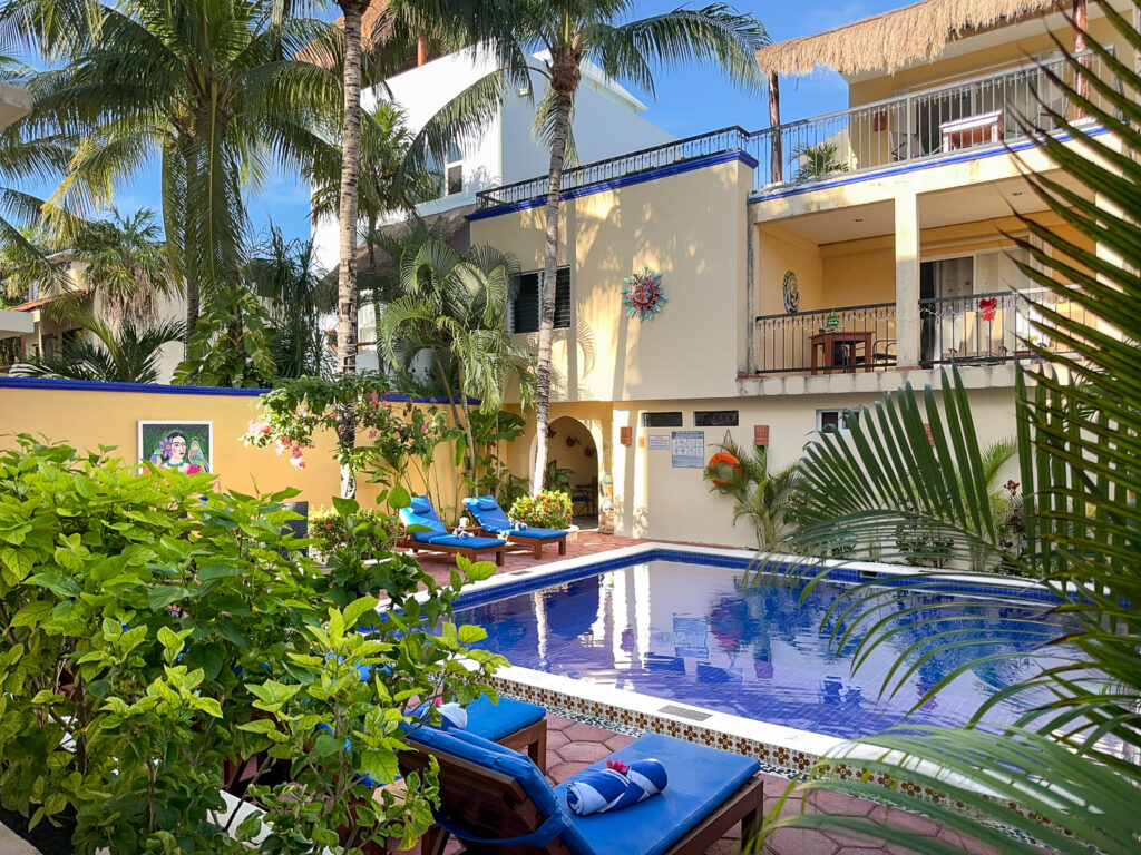 Pool surrounded by tropical greenery and yellow buildings with balconies and blue trim.