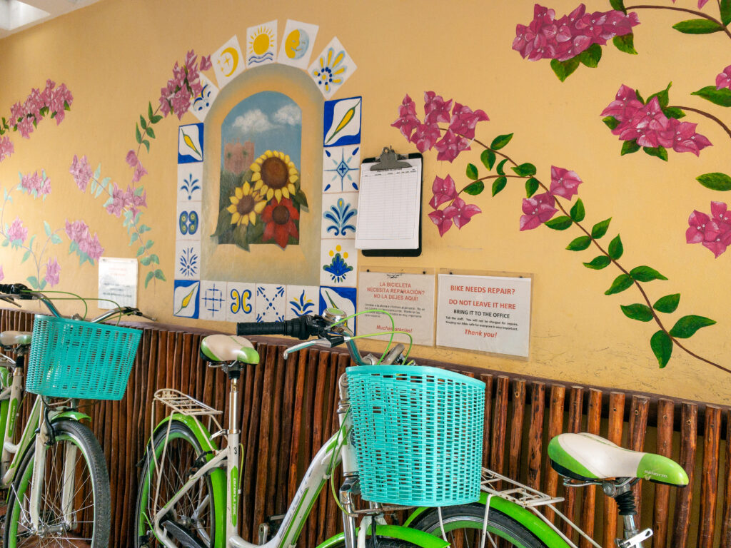 Bicycles with turquoise baskets beside a yellow wall painted with flowers and a decorative window.