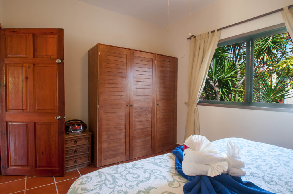 Patterned bedspread with folded towel decoration facing wooden closet doors and a curtained window.