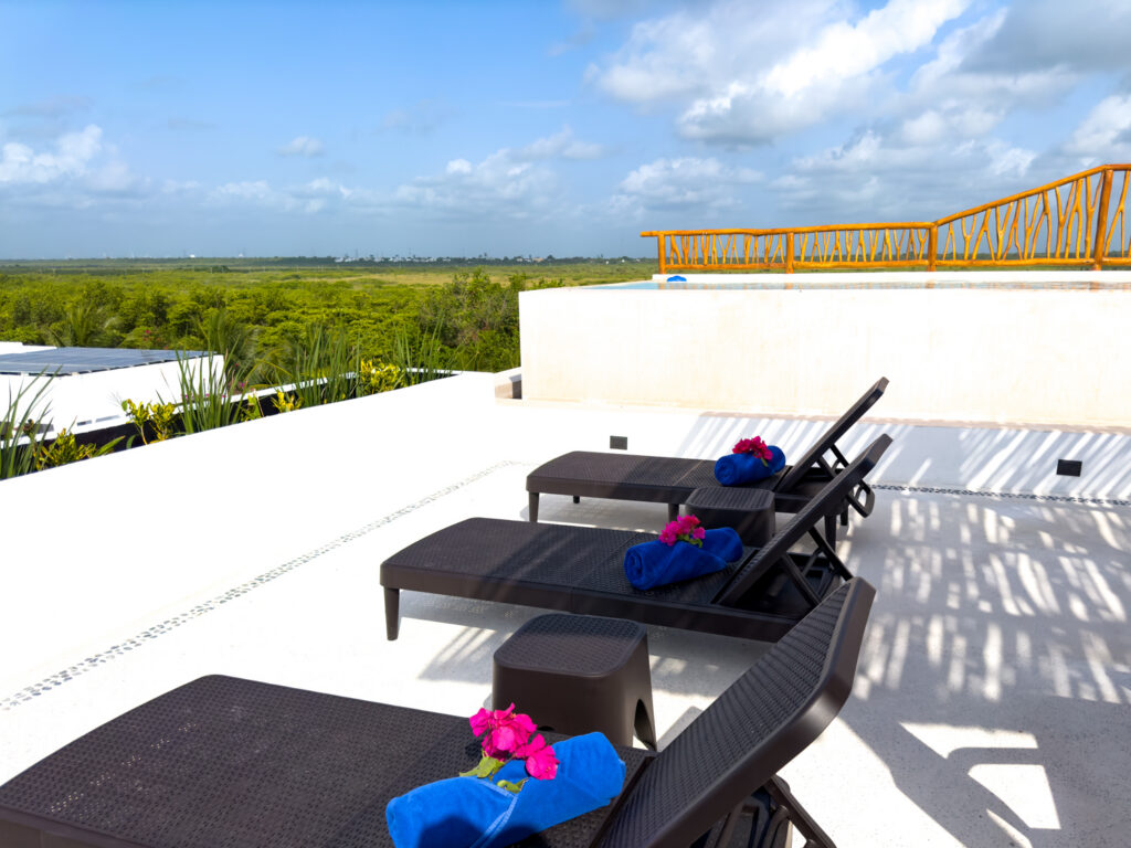 Dark lounge chairs with rolled blue towels on a white rooftop terrace beneath scattered clouds.