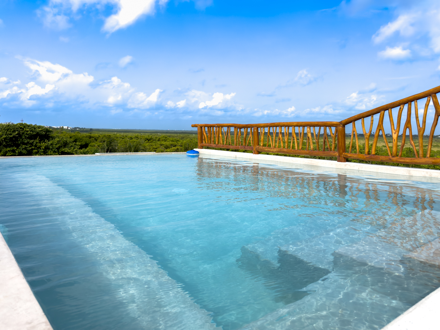 Blue rooftop pool with shallow steps and a wooden railing overlooking a green horizon.