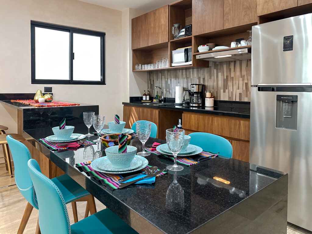 Dining table set with blue dishes beside wooden kitchen cabinets and a stainless steel refrigerator.
