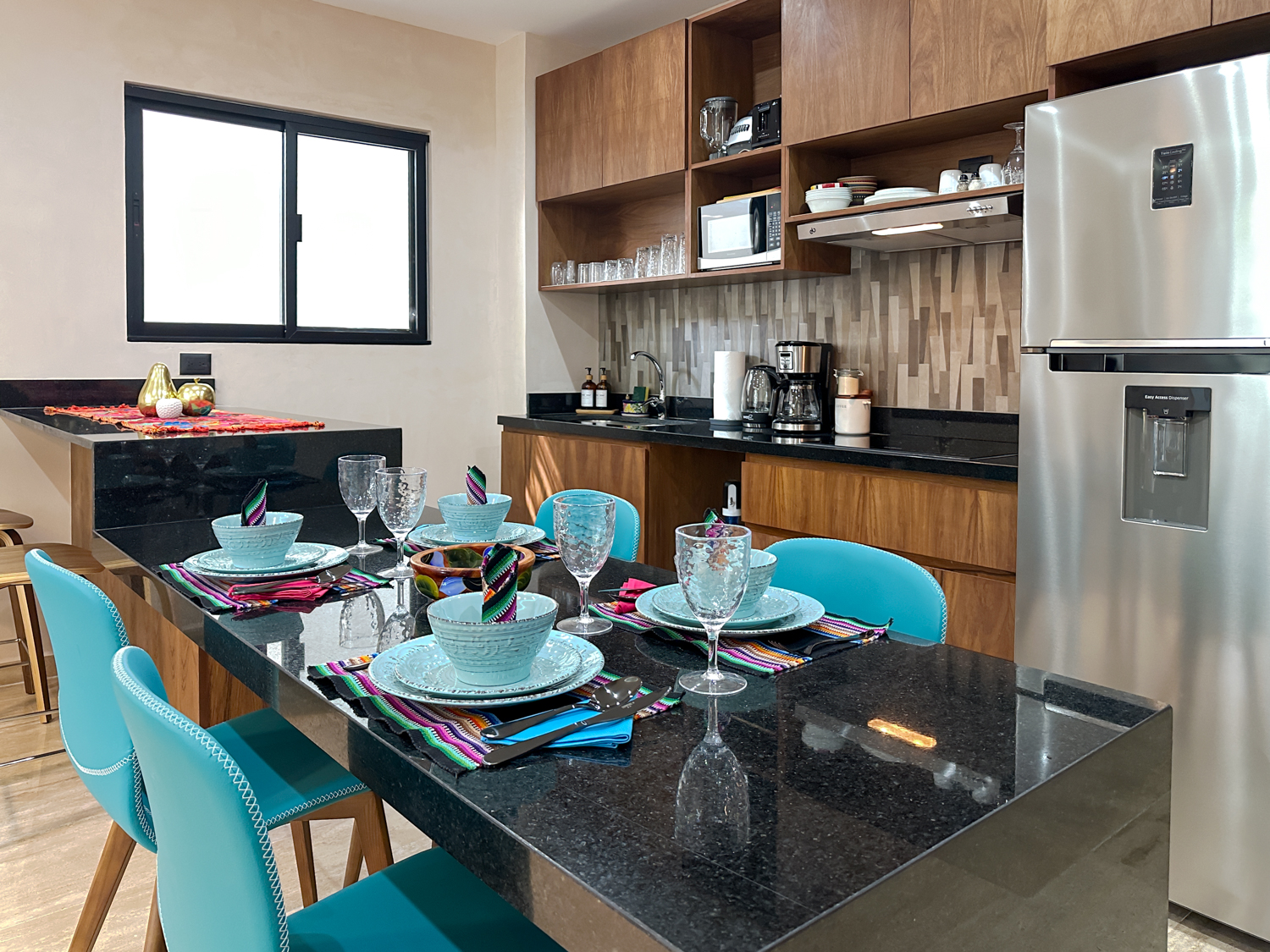 Dining table set with blue dishes beside wooden kitchen cabinets and a stainless steel refrigerator.