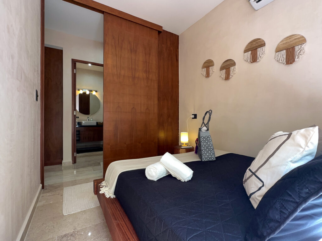 Dark-covered bed with rolled towels beside wooden storage and an open bathroom doorway.