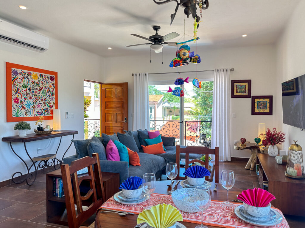 Round table set with colorful dishes and folded napkins in front of a gray sectional sofa.