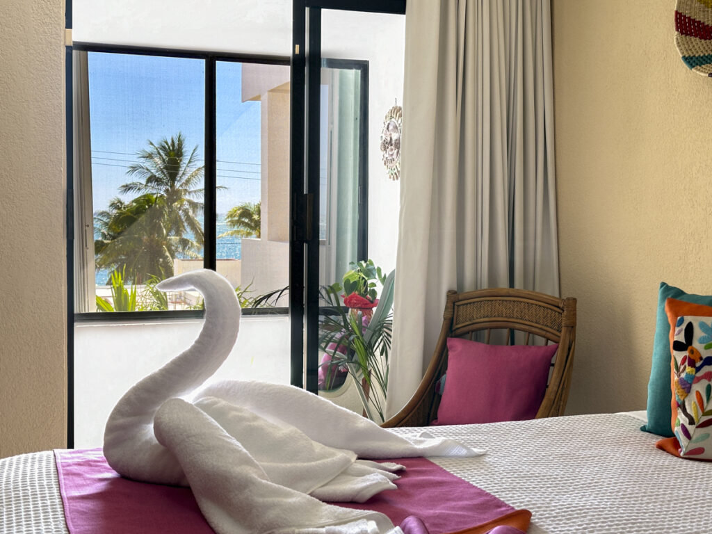 Towel swan on a bed beside a window overlooking palms and a wicker chair.