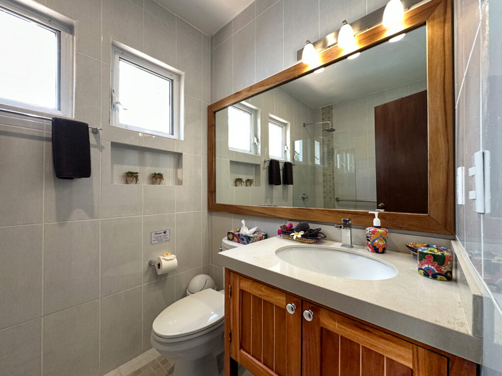 Wide bathroom mirror above a stone counter and wooden vanity beside a toilet and high window.