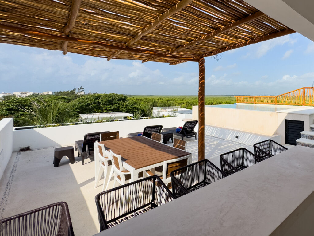Dining table and chairs beneath a wooden shade canopy beside rooftop lounge seating.