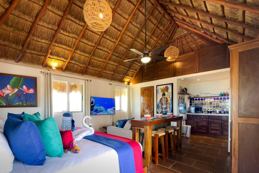 Bed, dining bar, and kitchen beneath a high thatched ceiling with woven pendant lights.
