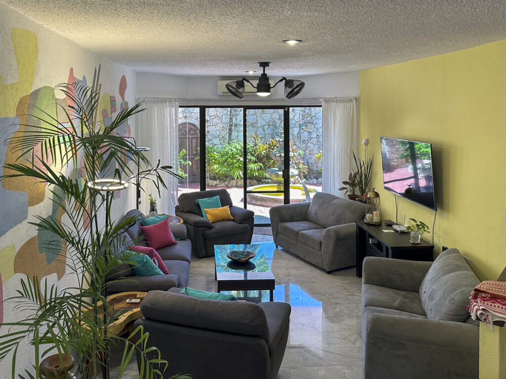 Long living room with gray seating, glass table, television, and abstract mural beside a yellow wall.