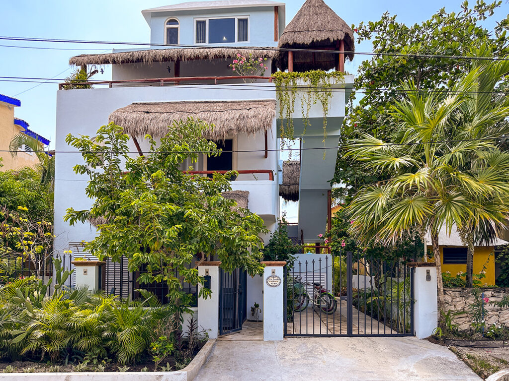White multistory building with balconies and thatched awnings behind trees and a gated entrance.