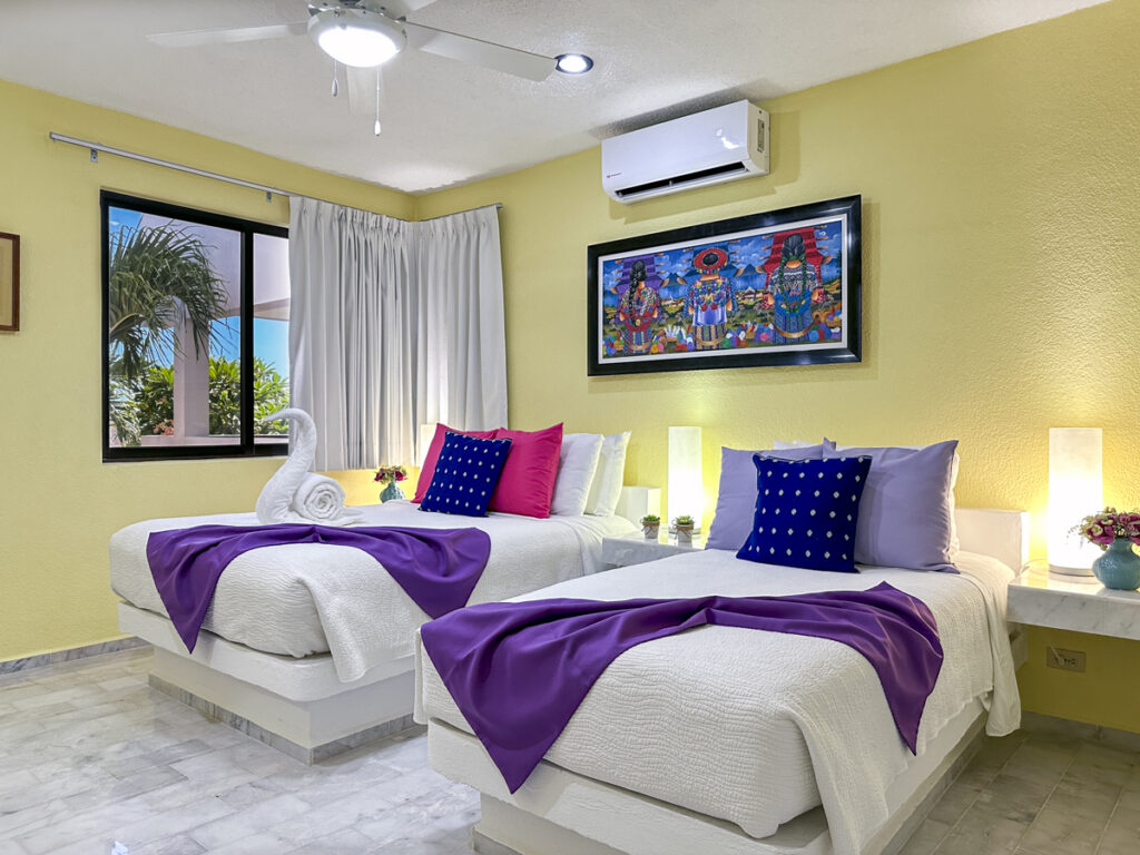 Two white beds with purple throws and blue cushions beneath colorful artwork.