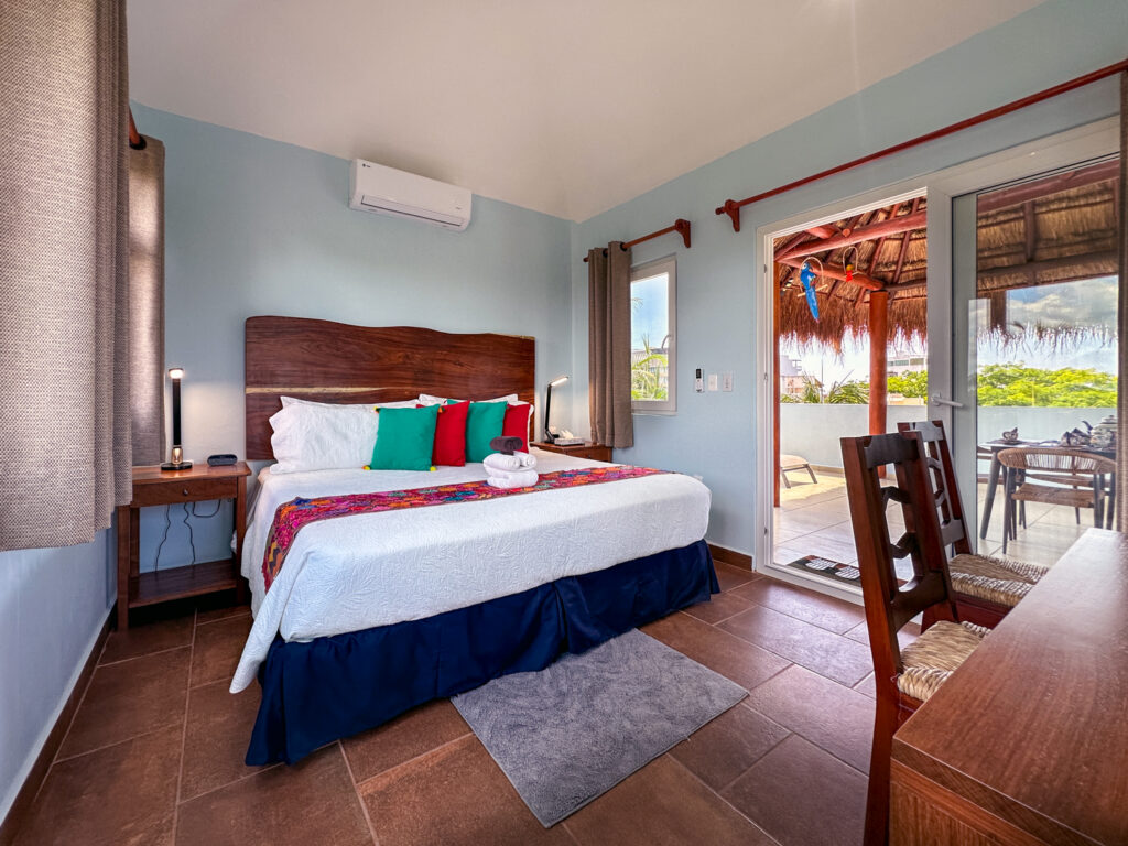 Bed with floral runner and colorful pillows beside sliding doors opening onto a thatched terrace.