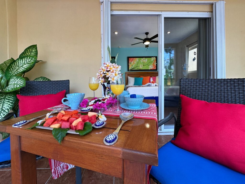 Balcony table with fruit and flowers between cushioned chairs outside the bedroom doors.
