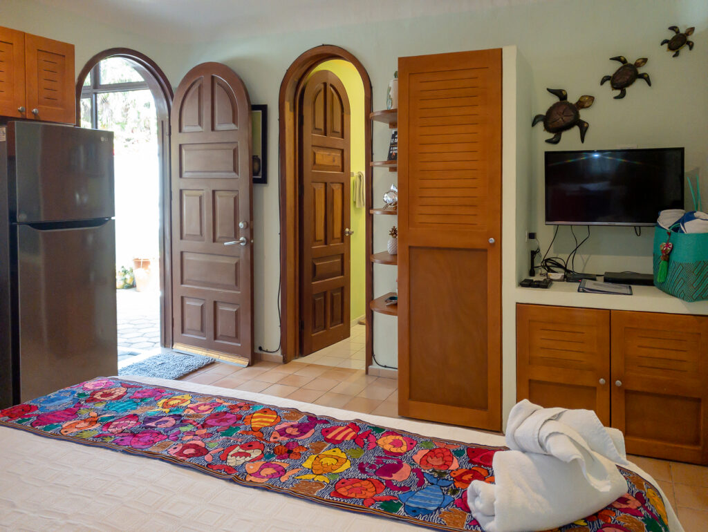 Bed with a floral runner facing an arched wooden doorway, refrigerator, storage cabinet, and television.