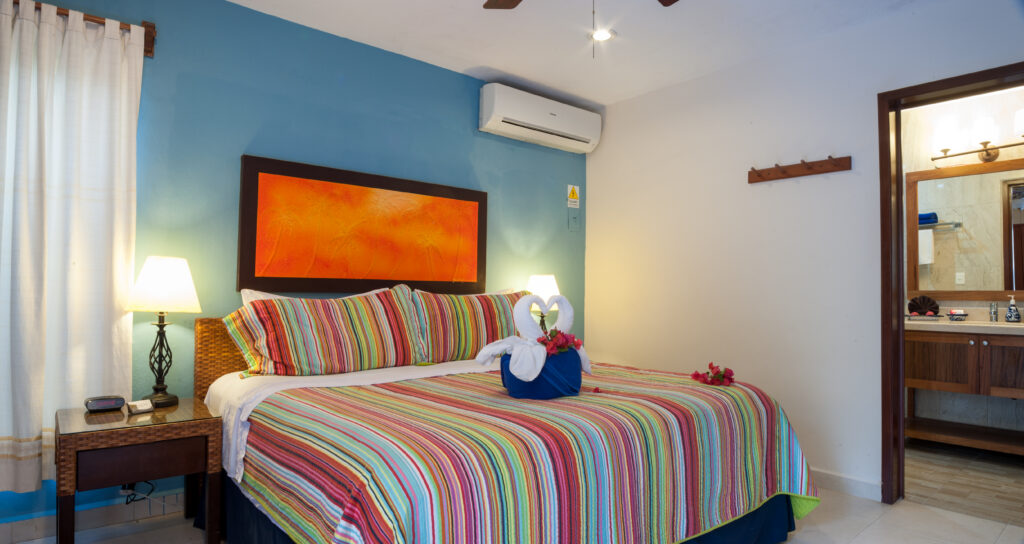 Bed with striped bedding beneath orange artwork beside an open bathroom doorway.