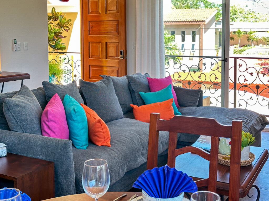 Gray sectional with bright pink, turquoise, and orange cushions beside balcony doors and iron railings.