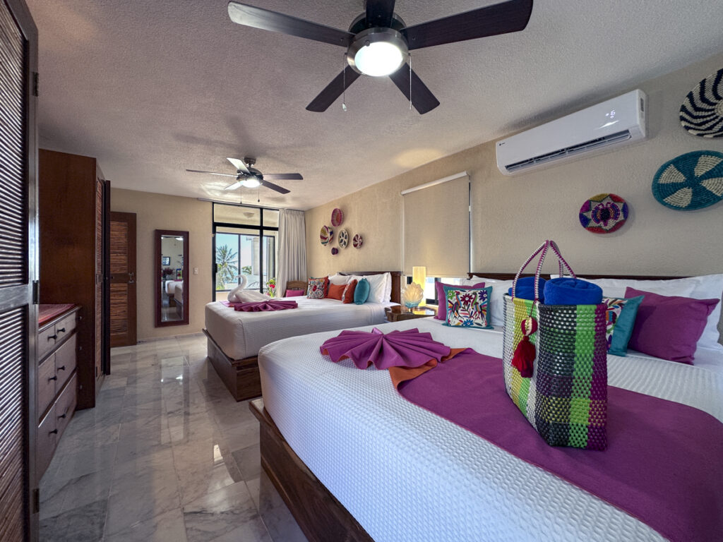 Long bedroom with two large beds, colorful cushions, ceiling fans, and wall decorations.