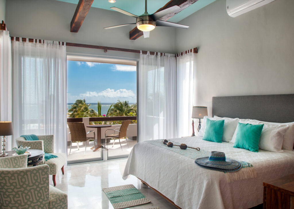 White bed with turquoise pillows beside sliding balcony doors overlooking palms and the coast.