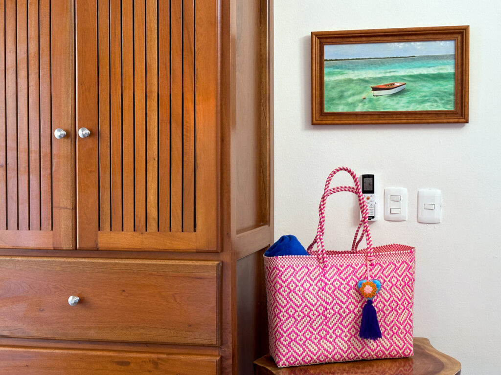Wooden wardrobe beside a pink woven bag on a small table beneath a framed seascape.