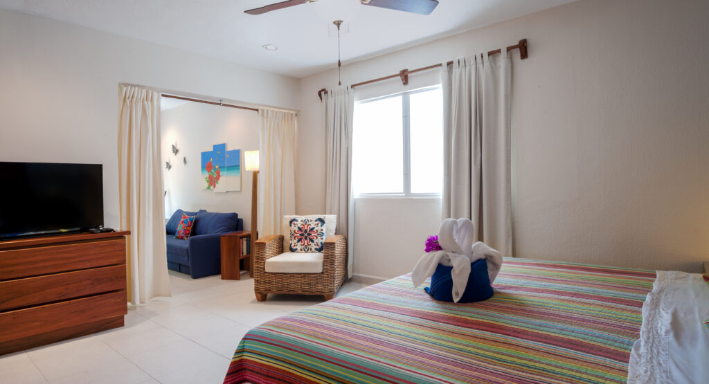 Bedroom with a striped bedspread, television, wicker chair, and doorway leading to the blue sofa.