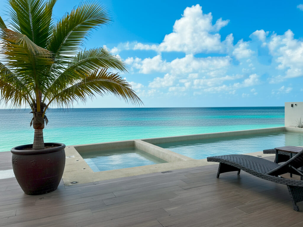 Potted palm and lounge chairs beside a long pool overlooking turquoise Caribbean water.