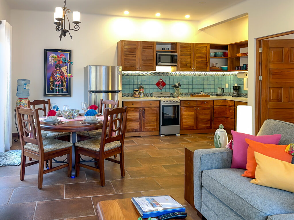 Dining table and sofa beside a kitchen with wooden cabinets, blue tiles, and stainless steel appliances.
